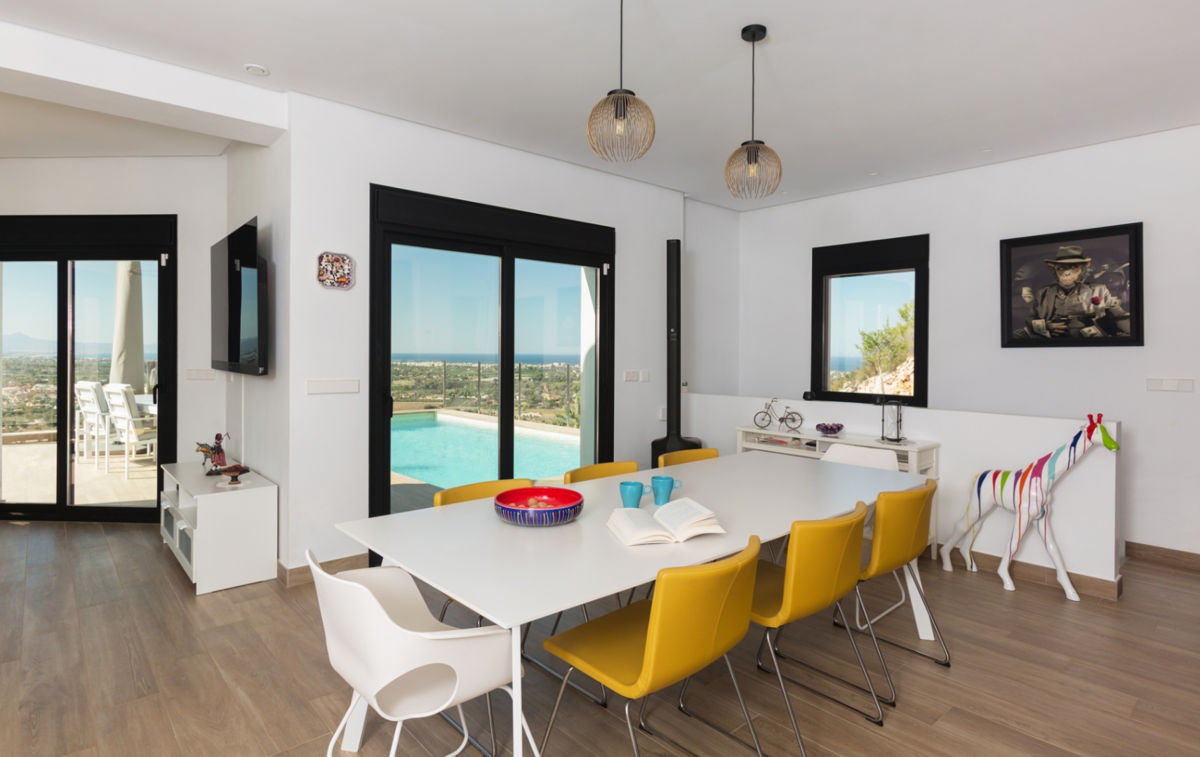 Moderne eetkamer met witte tafel en gele stoelen, houtlook tegelvloer en schuifpui naar het terras met zwembad en panoramisch uitzicht over de kustlijn.