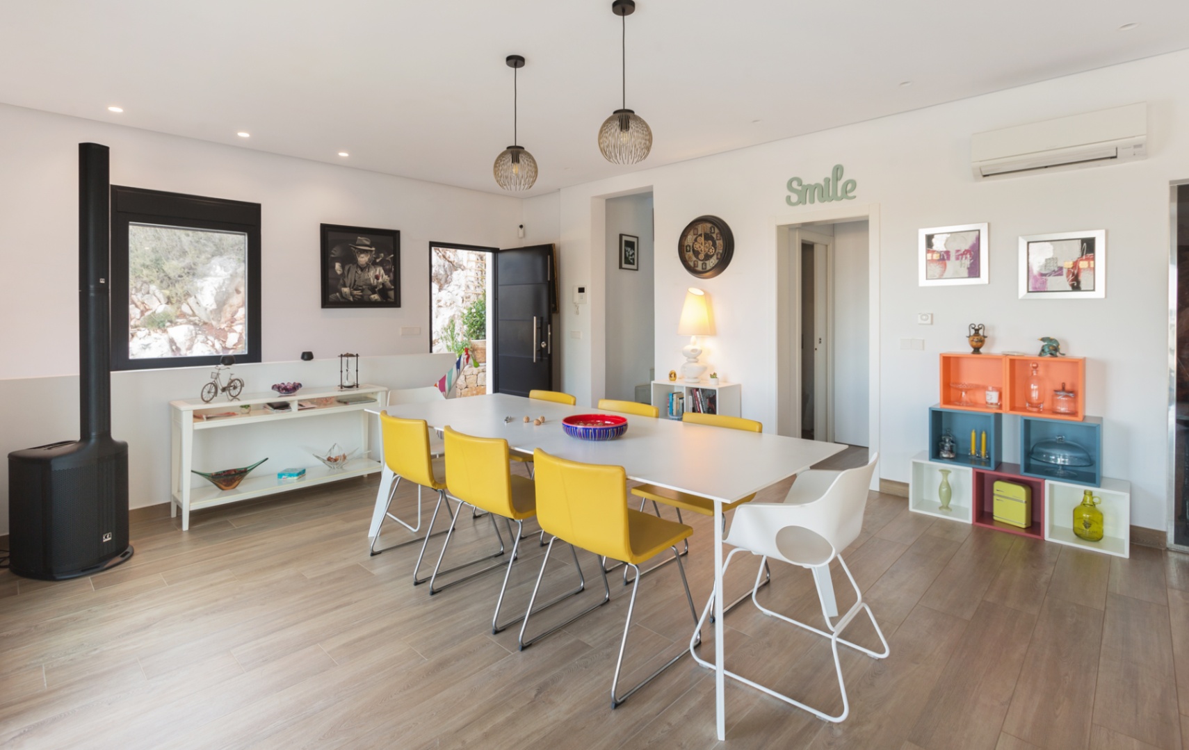 Moderne eetkamer met witte tafel, gele stoelen, houtlook vloer, een strakke zwarte houtkachel en kleurrijke modulaire open kasten.