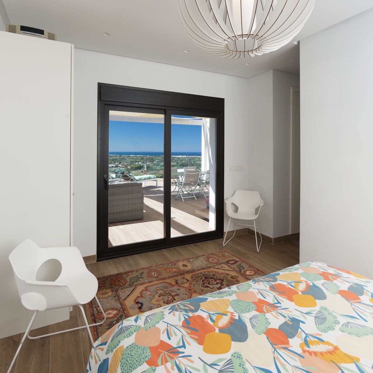 Moderne slaapkamer met een glazen schuifdeur naar een gemeubileerd terras met uitzicht op zee. Voorzien van een sculpturale hanglamp en witte designstoelen.
