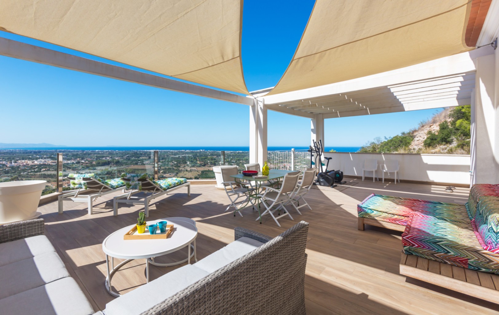 Ruim dakterras met houtlook vloertegels, schaduwdoeken en panoramisch uitzicht op zee. Voorzien van een eethoek, loungesets en ligbedden onder een witte pergola-constructie.
