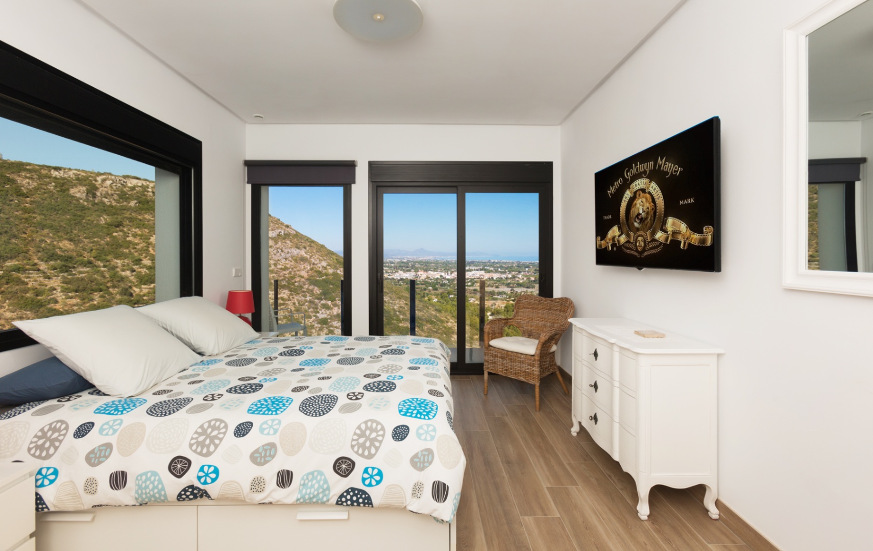 Slaapkamer met panoramisch uitzicht op bergen en zee, grote zwart omlijste ramen, houtlook vloer, wit dressoir en een aan de muur gemonteerde televisie.
