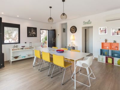 Lichte eetkamer met een lange witte tafel, zes gele en één witte stoel, verlicht door moderne hanglampen. Voorzien van lichte hout-look vloer en eclectische decoratie.