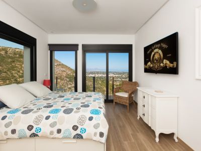 Lichte slaapkamer met designbed, houten vloer en grote zwartomlijste ramen die panoramisch uitzicht bieden op de bergen en de kust. Inclusief witte ladekast en tv.