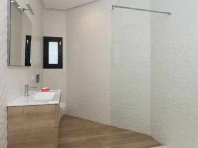 Moderne badkamer met zwevend houten wastafelmeubel, witte wastafel, verlichte spiegel en inloopdouche met glaswand. Opvallende witte tegels met geometrische textuur en houtlook vloertegels.