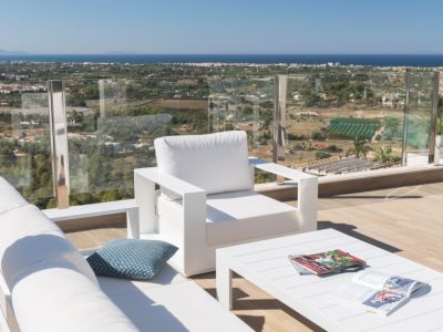 Moderne witte zithoek buiten op een houten terras, met een glazen balustrade die een panoramisch uitzicht biedt op de kustlijn en de Middellandse Zee onder een heldere hemel.