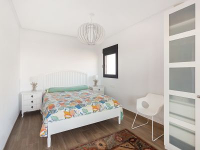 Slaapkamer met wit lattenbodem hoofdbord, kleurrijk dekbed en houtlook tegelvloer. Voorzien van witte nachtkastjes met lampen, een raam en een kledingkast met matglazen panelen.