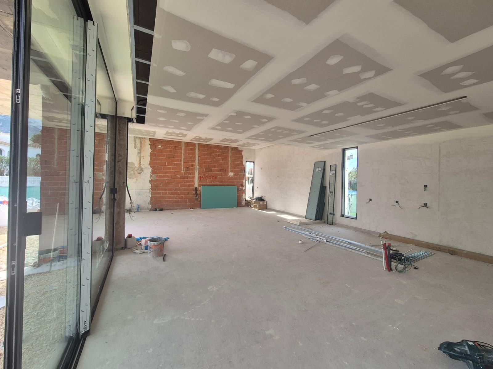 Ruime woonkamer in aanbouw met gipsplaten plafond voor inbouwspots, een bakstenen muur en grote glazen schuifdeuren van vloer tot plafond.