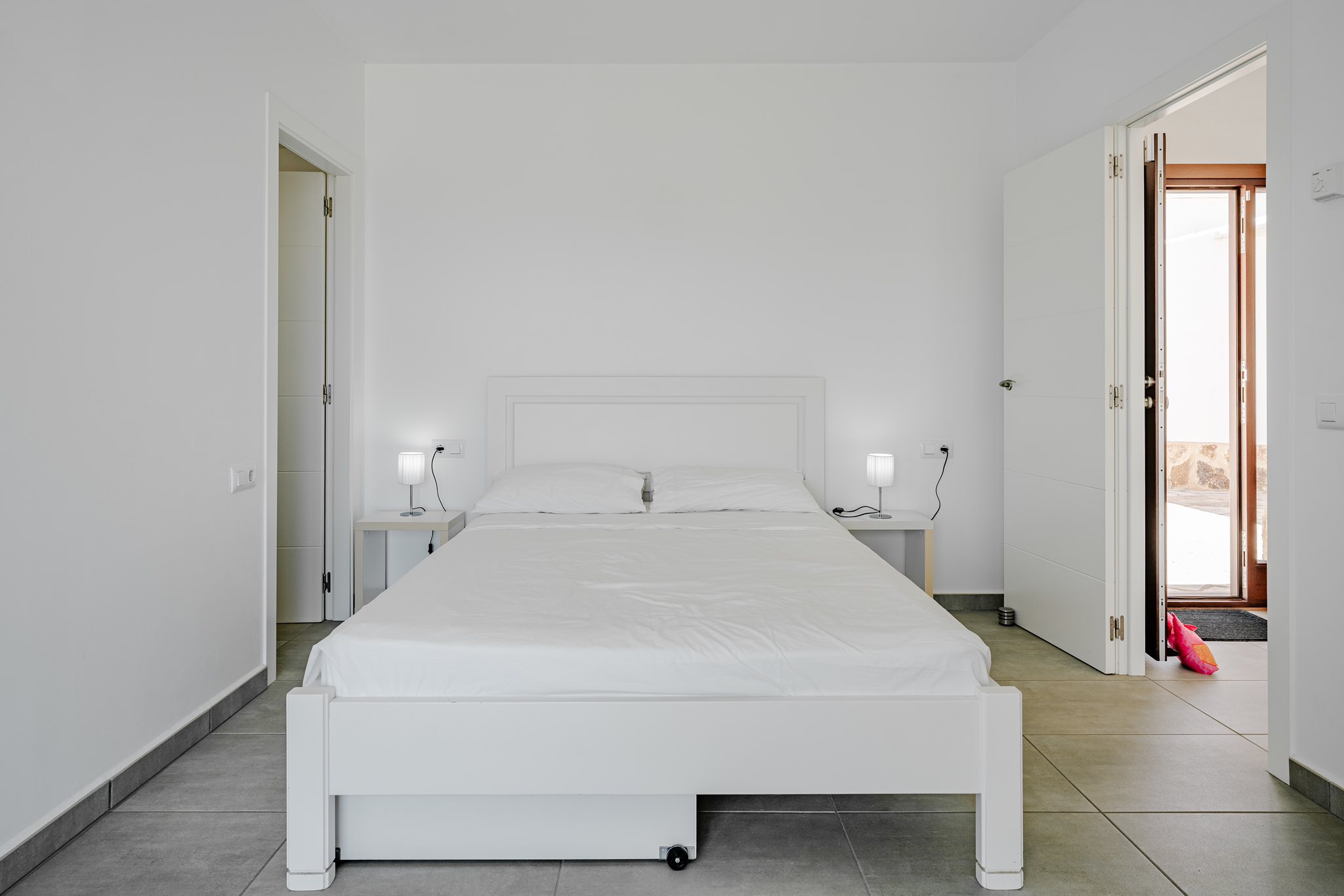 Minimalistische slaapkamer met een wit houten bed, bijpassende nachtkastjes en grijze tegelvloer. Open deuren bieden toegang tot de aangrenzende kamers en buiten.