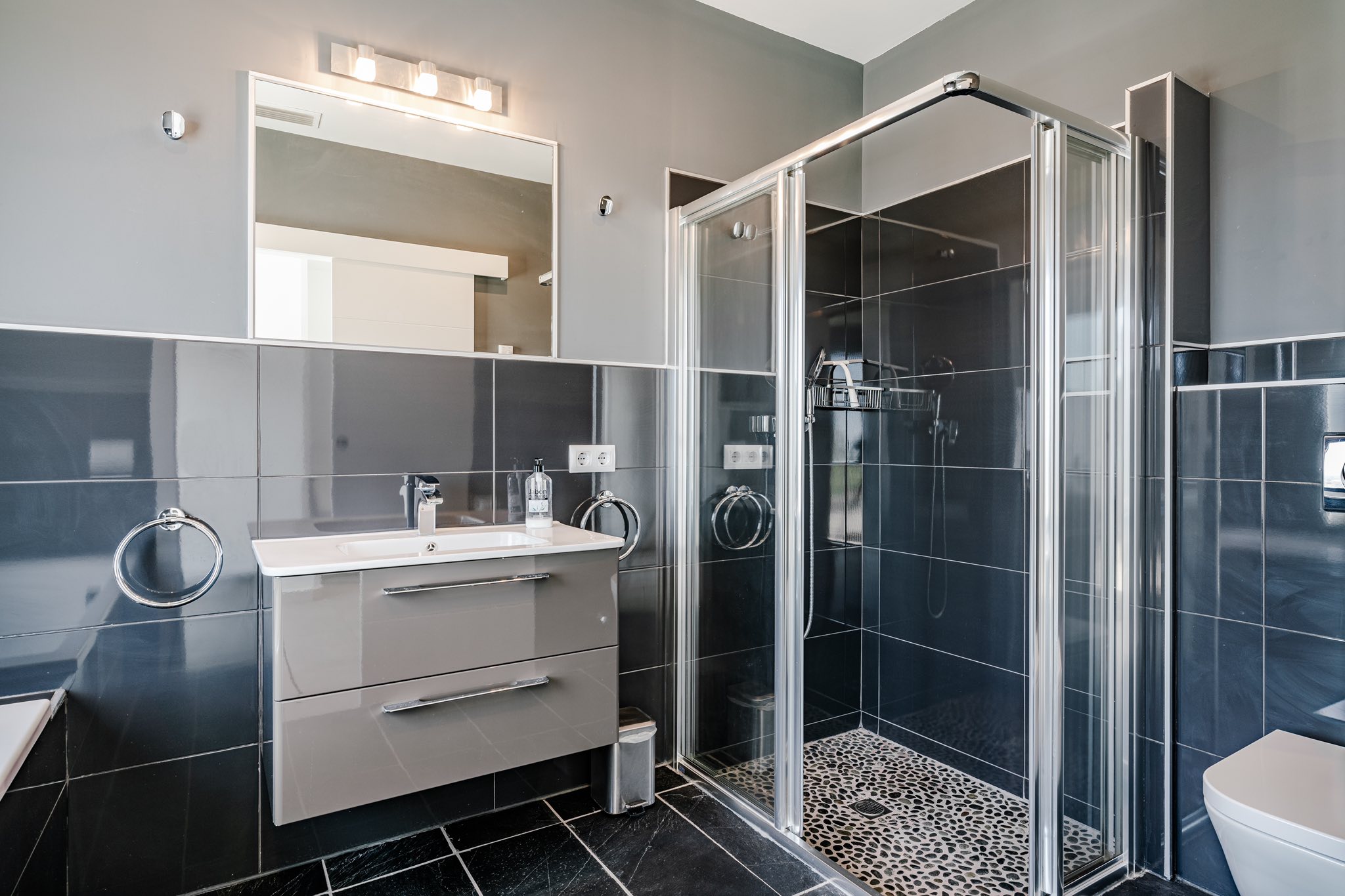 Moderne badkamer met antracietkleurige glanzende tegels, een zwevend wastafelmeubel en een glazen douchecabine met decoratieve kiezelvloer.