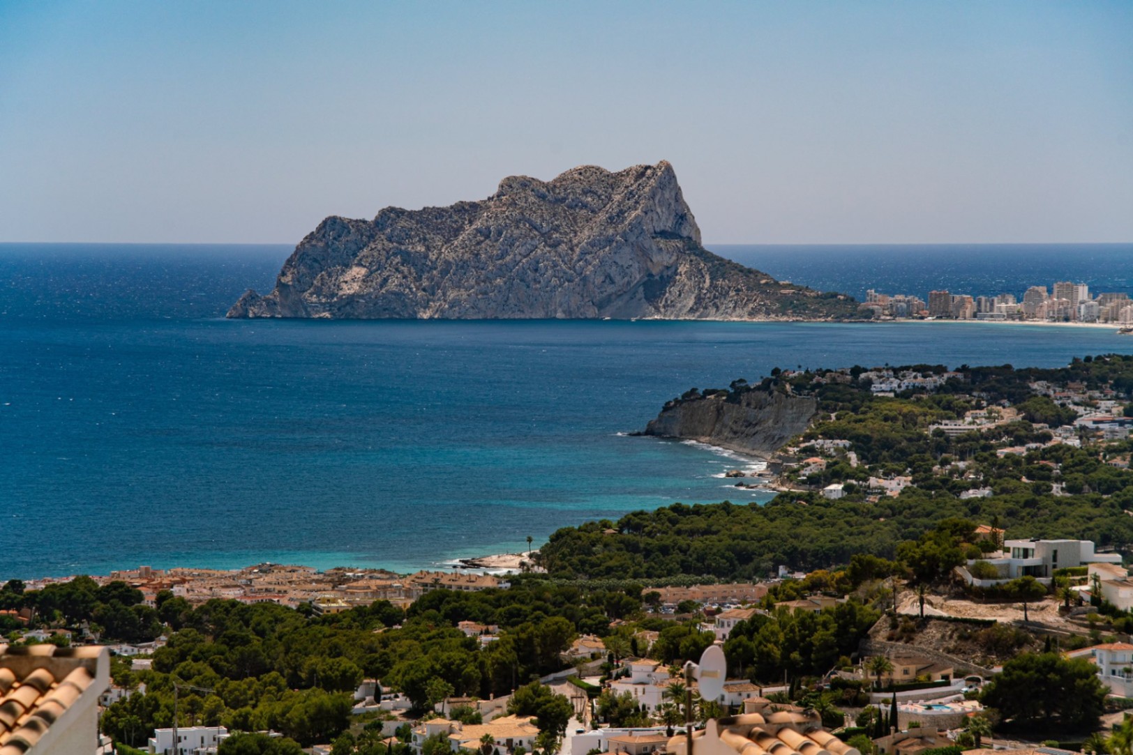 Verhoogd panoramisch uitzicht op de Middellandse Zeekust met de Peñón de Ifach, turquoise wateren en een woonwijk te midden van dichte dennenbossen.