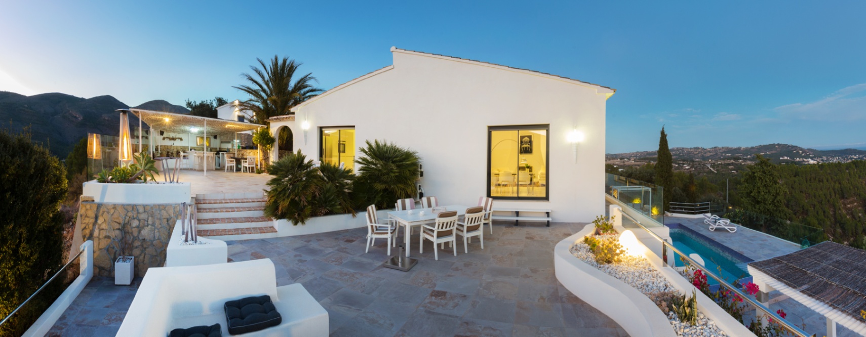 Groot stenen terras bij schemering met eethoek, geïntegreerde plantenbakken en panoramisch uitzicht over de vallei naast een witte mediterrane villa.