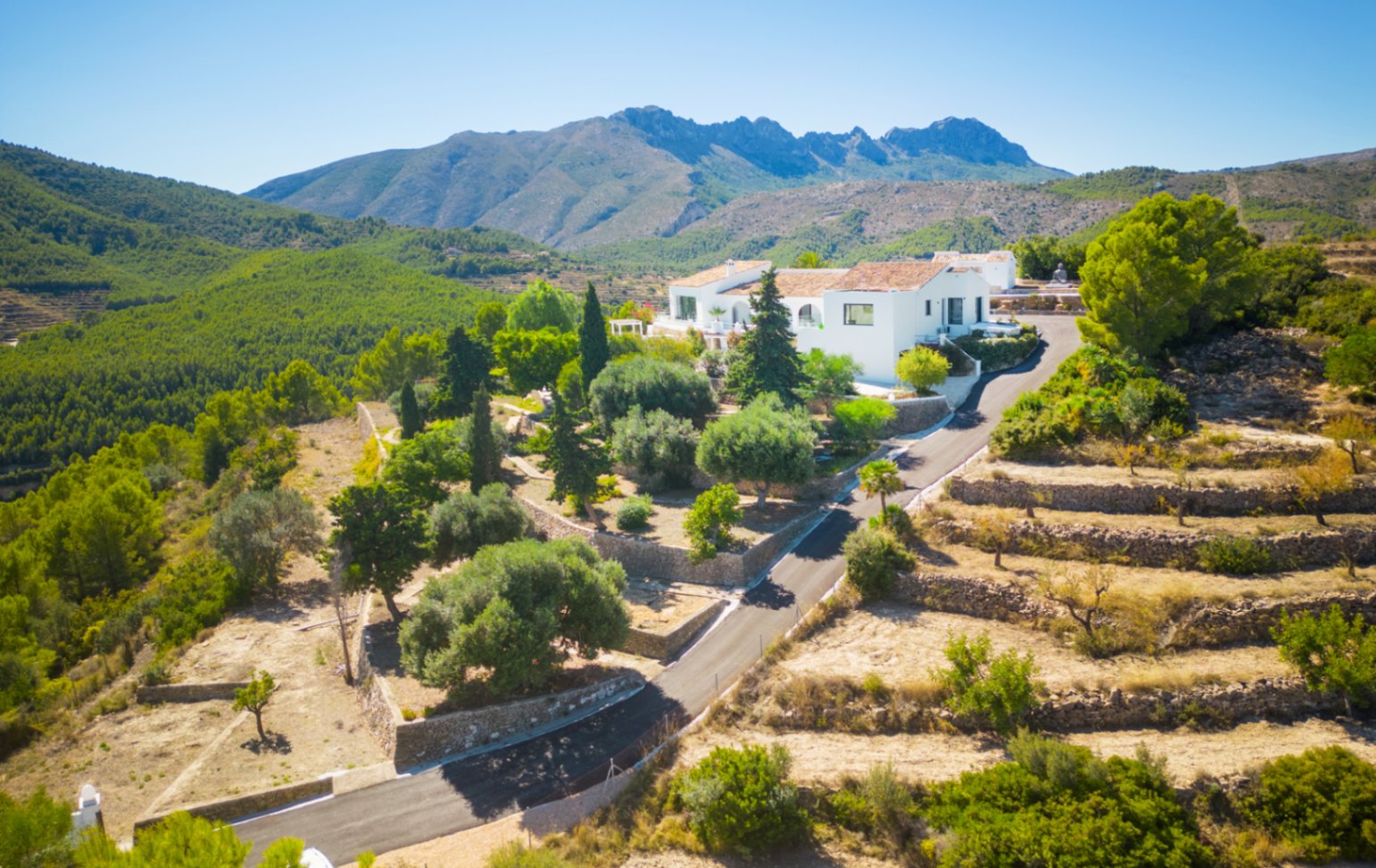 Luchtfoto van een witte villa in een bergachtig landschap met stenen terrassen, mediterrane tuinen en panoramisch uitzicht op het omliggende dennenbos.