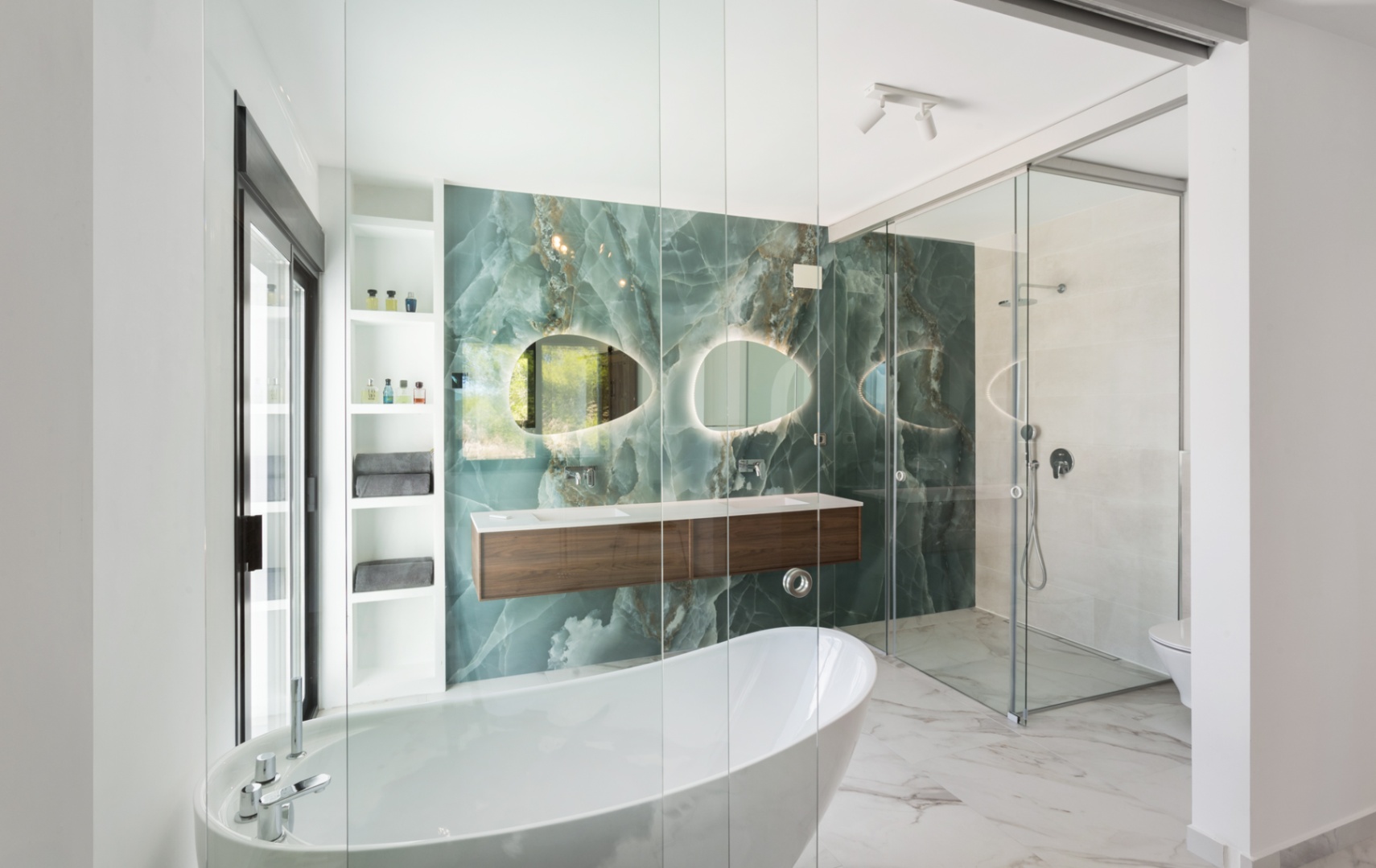 Moderne badkamer met een groene onyx wand, vrijstaand ligbad en zwevend houten wastafelmeubel. Voorzien van organische verlichte spiegels en een glazen inloopdouche.