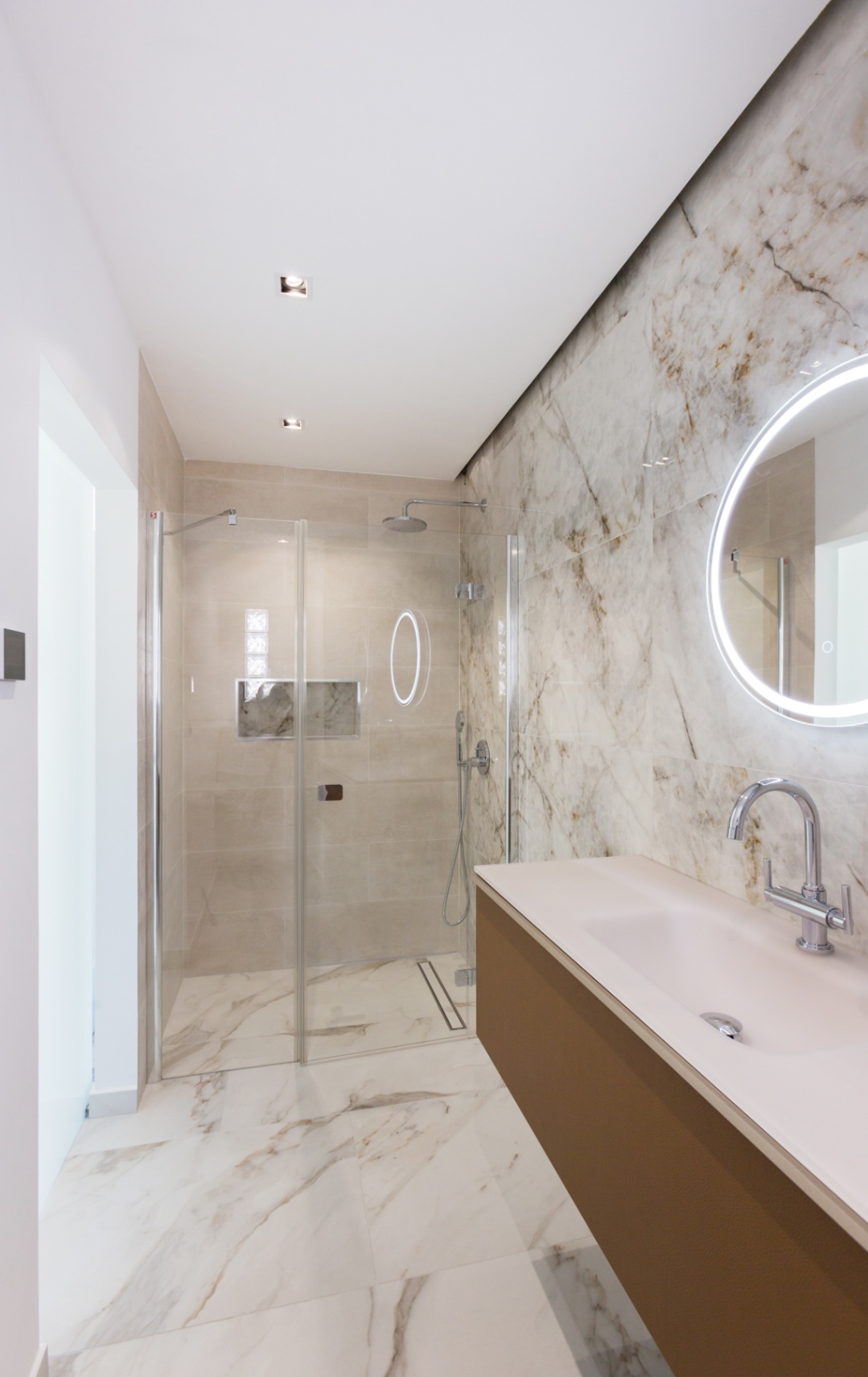 Moderne badkamer met inloopdouche van glas, tegels met marmerlook, een zwevend wastafelmeubel en een ronde spiegel met geïntegreerde LED-verlichting.