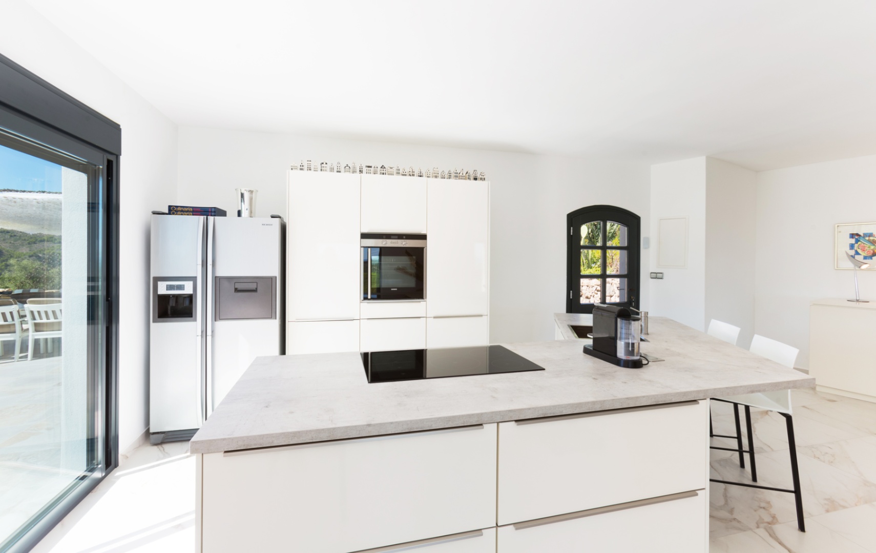 Moderne keuken met kookeiland in steenlook, inductiekookplaat, witte kasten en een Amerikaanse koelkast. Voorzien van een lichte marmeren vloer.