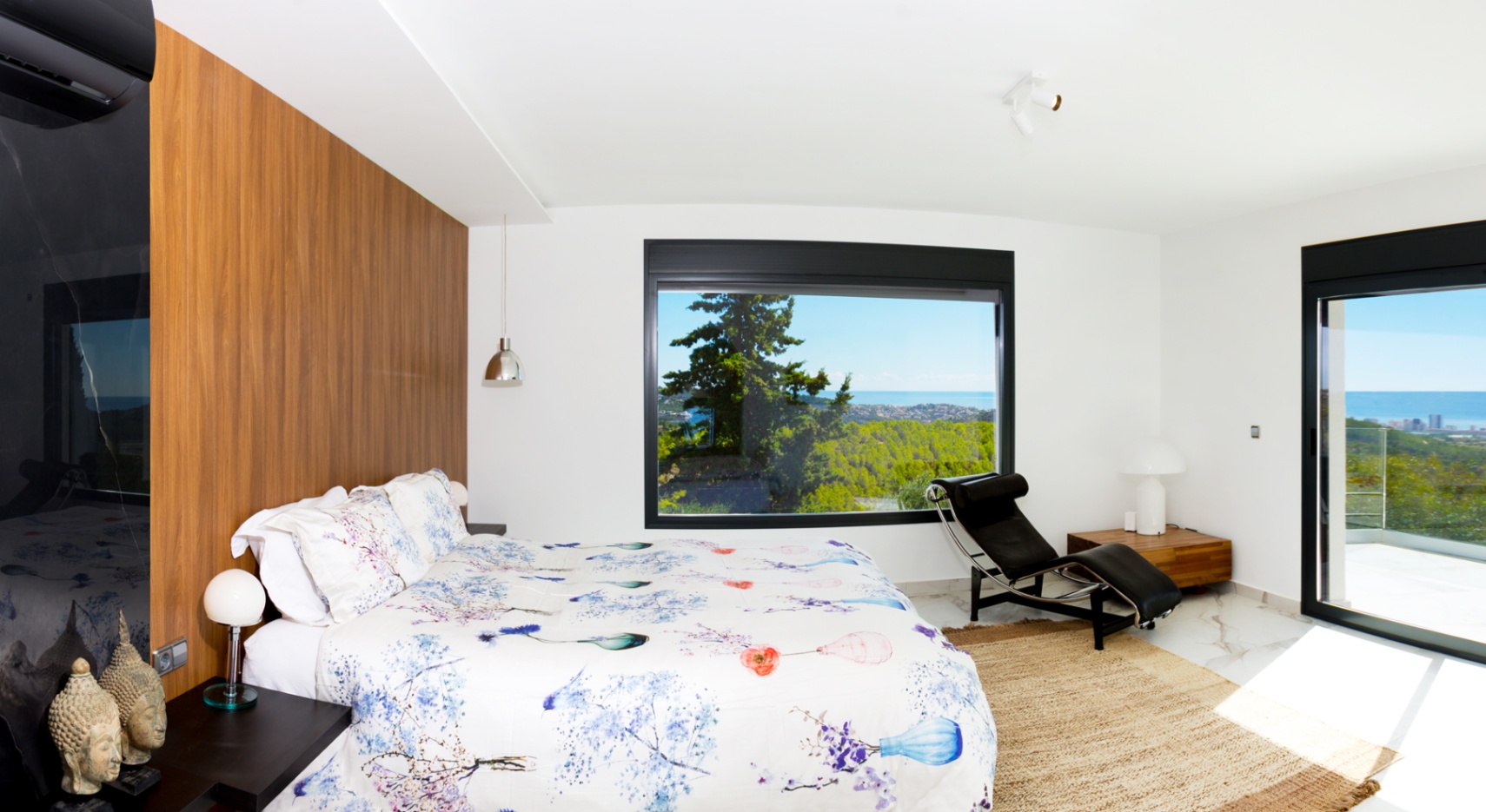 Moderne slaapkamer met houten accentwand, marmeren vloer en een groot panoramisch raam met uitzicht op zee en het bos. Inclusief een design chaise longue.