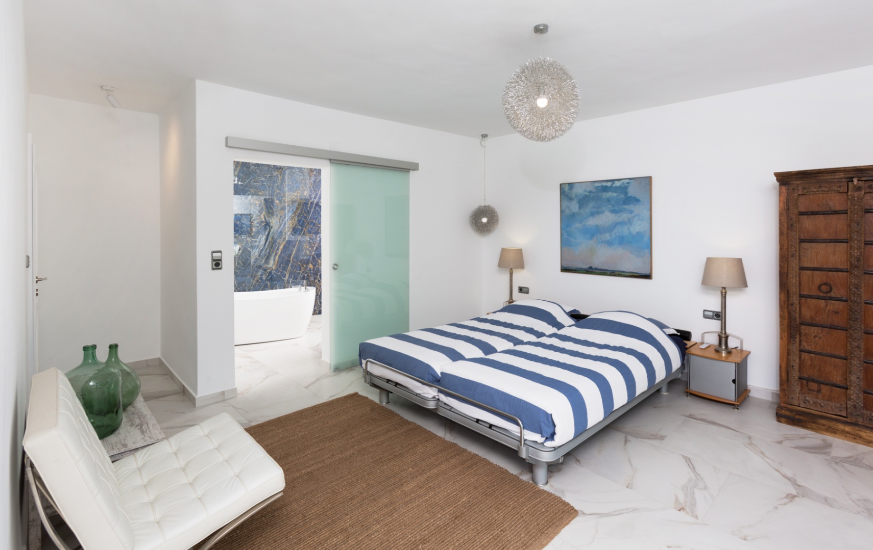 Moderne slaapkamer met marmerlook vloer, blauw gestreept beddengoed en glazen schuifdeur naar de badkamer met vrijstaand bad en blauwe natuurstenen wand.