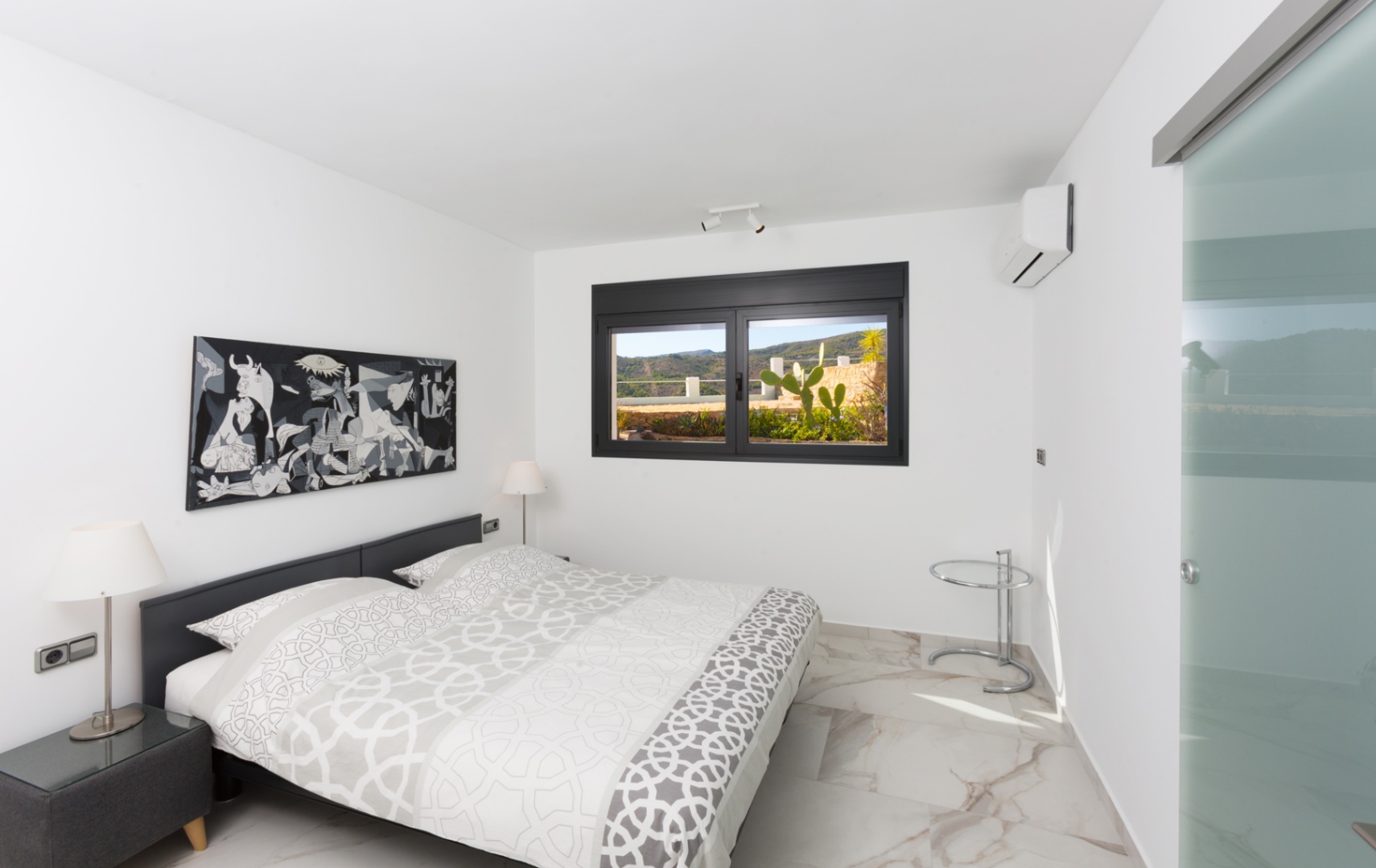 Moderne slaapkamer met marmerlook vloertegels, een monochroom kunstwerk en een zwart raamkozijn met uitzicht op een terras met cactussen en heuvels.