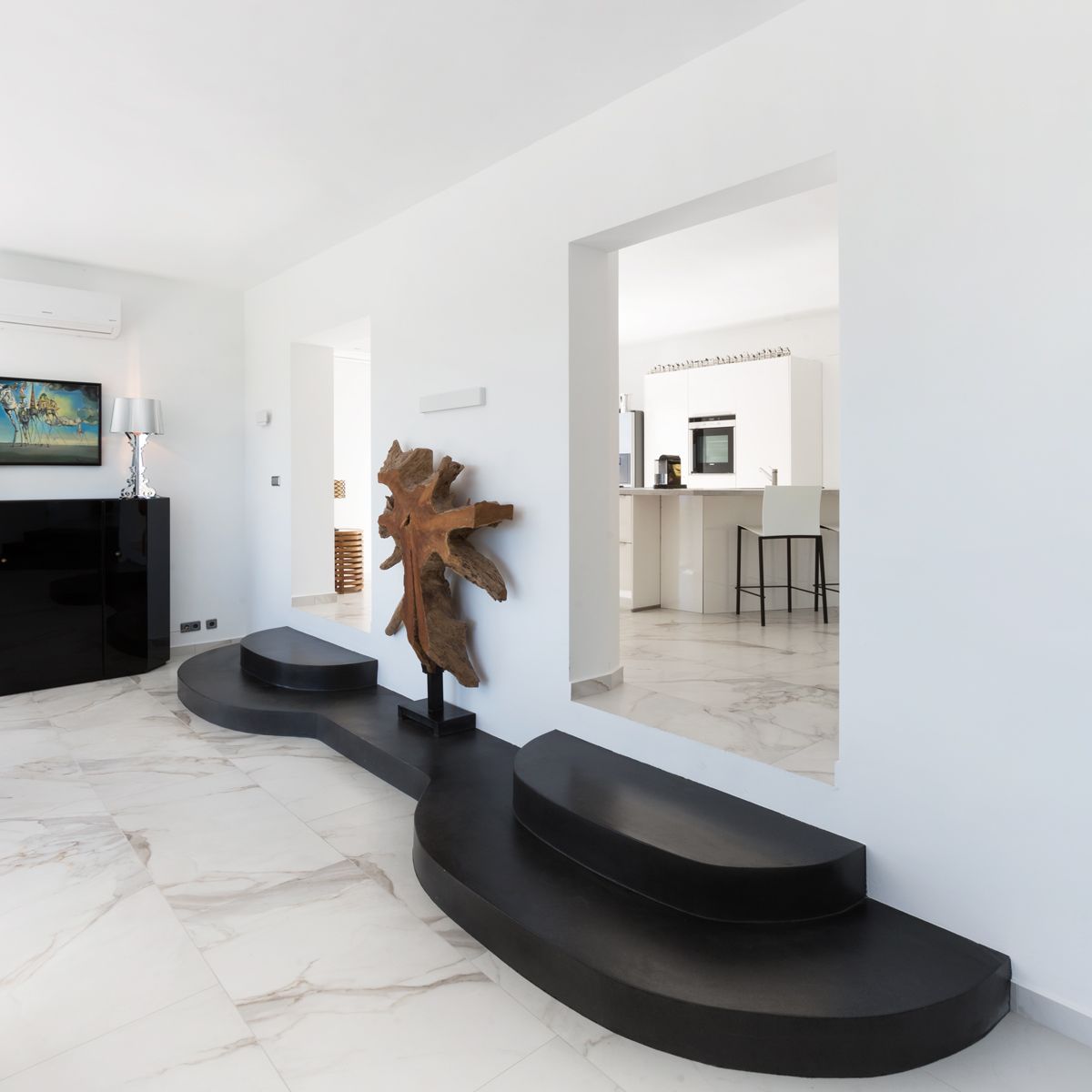 Moderne woonkamer met witte marmeren vloer en een uniek zwart gebogen platform. Een natuurlijke houten sculptuur siert de doorgang naar de strakke witte keuken.