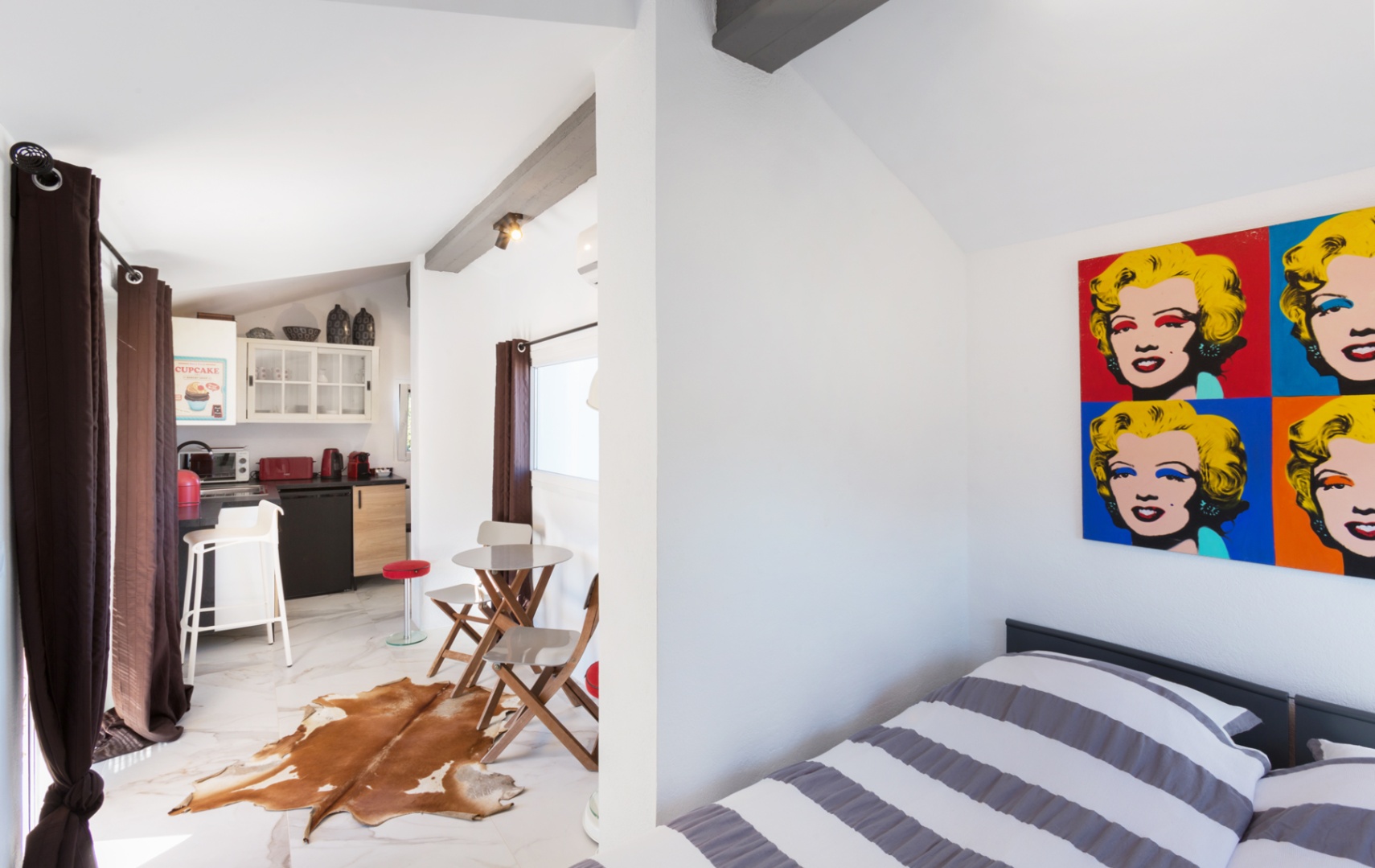 Studio met pop-art decor, donkere balken en marmerlook vloer. Beschikt over een compacte kitchenette met rode apparatuur en een eethoek met een koeienhuid tapijt.