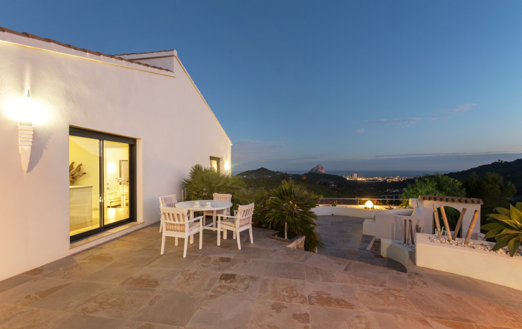 Terras met natuurstenen vloer, witte houten eethoek en panoramisch uitzicht op de kust van Calpe en de Peñón de Ifach tijdens de schemering.