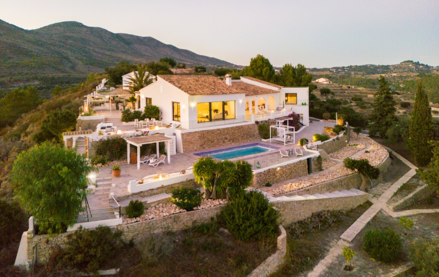 Verhoogd zicht op een mediterrane villa met witte gevels, terracotta daken en terrassen op verschillende niveaus met zwembad en natuurstenen muren.