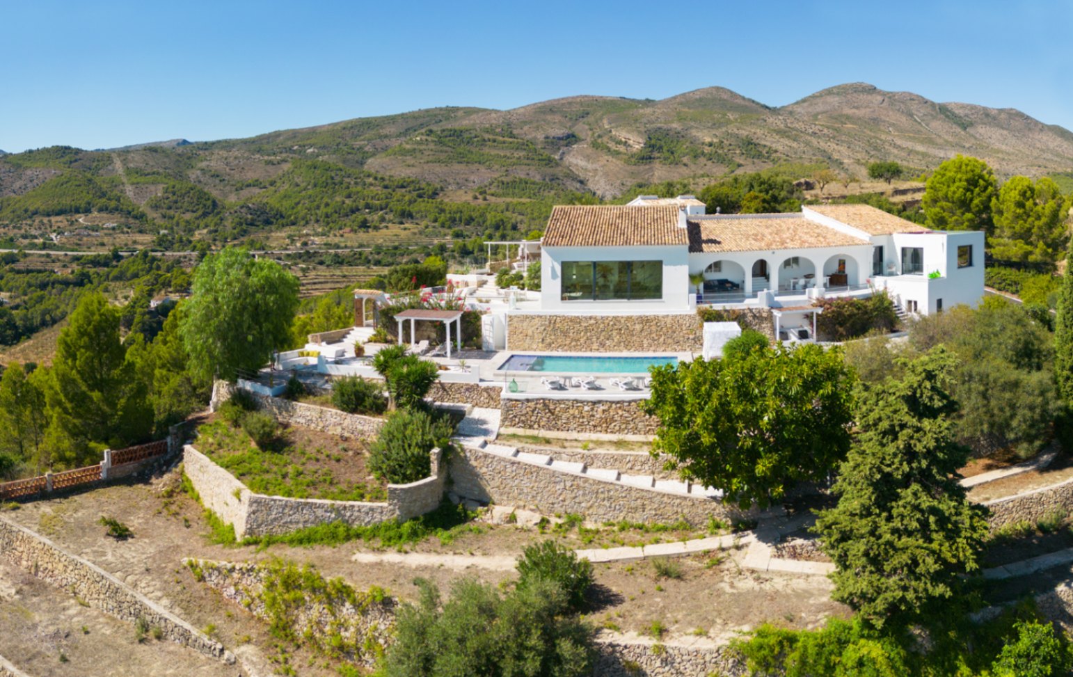 Verhoogd zicht op een witte mediterrane villa met terrastuinen van natuursteen, een privézwembad en een overdekt terras met bogen tegen een bergachtig decor.