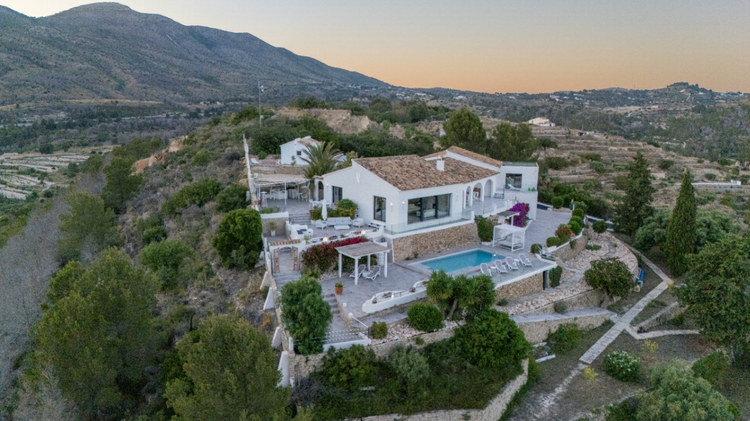 Verhoogd zicht op een witte mediterrane villa met terrasvormige stenen muren, een zwembad en diverse buitenruimtes tegen een bergachtig landschap.