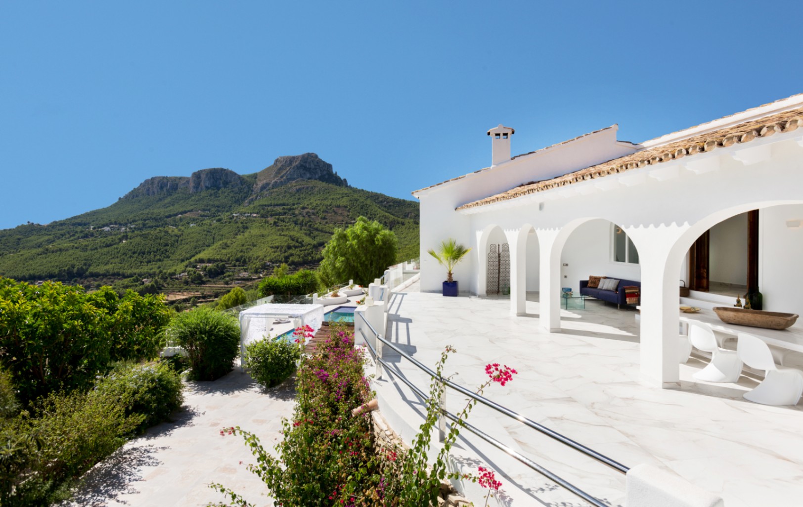 Witte mediterrane villa met een overdekt terras met bogen, marmeren vloer, buitenlounge en weids uitzicht op het groene berglandschap.