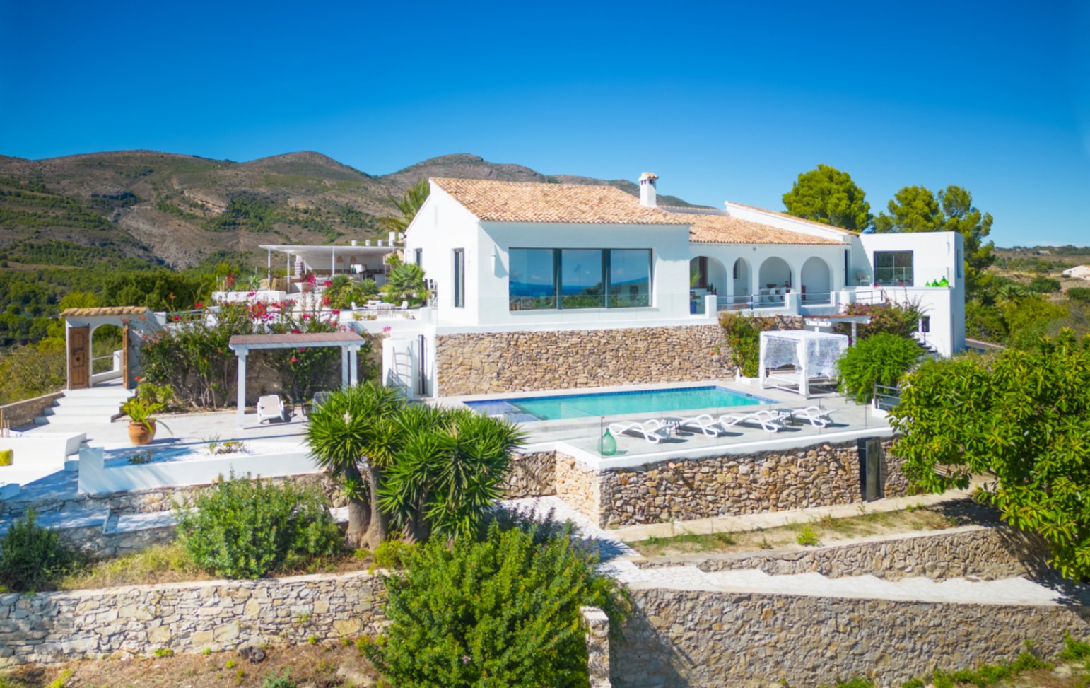 Witte mediterrane villa met zwembad, natuurstenen terrassen en een terracotta dak, gelegen in een bergachtig landschap.