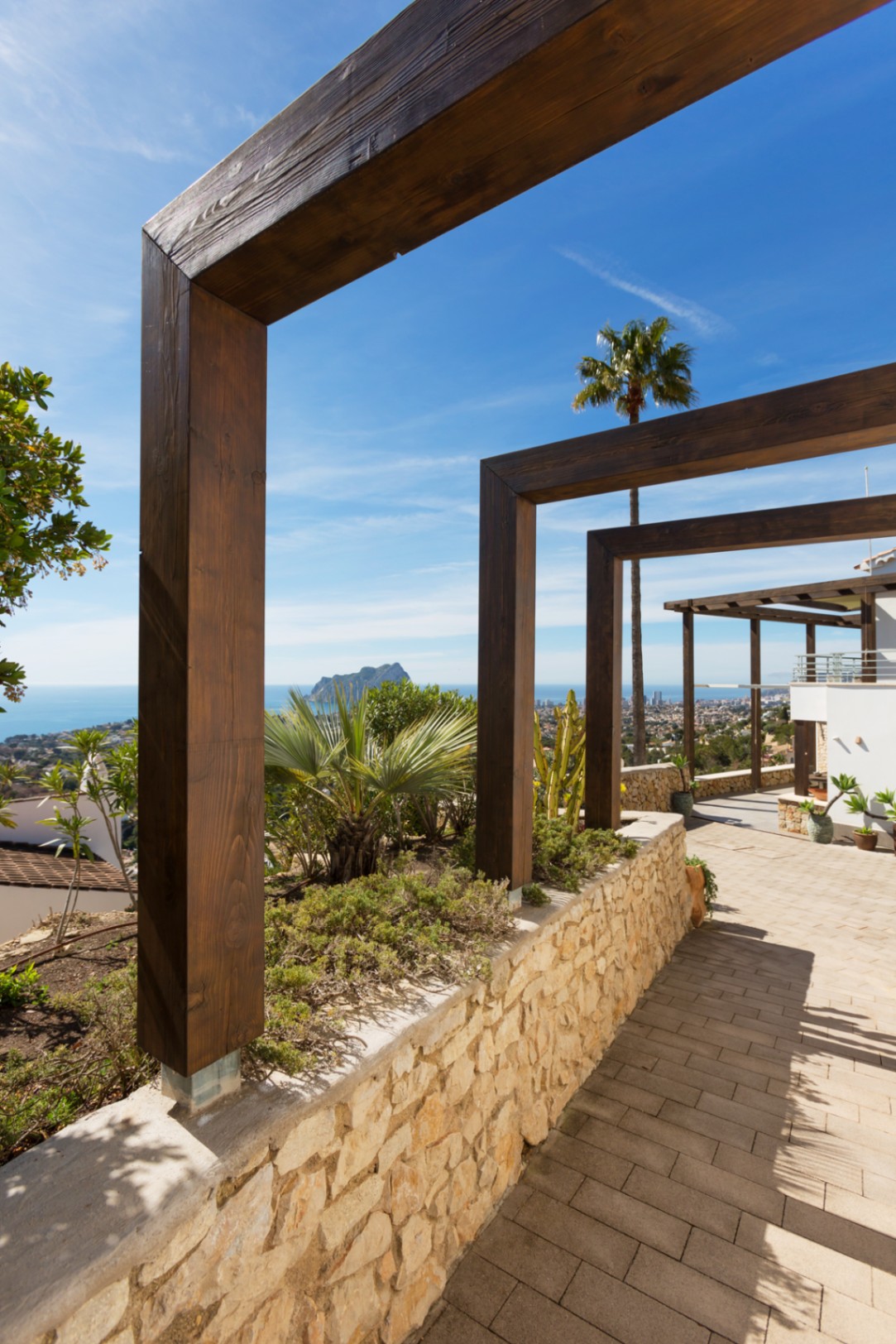 Buitenpad met donkerhouten pergola's, natuurstenen muur en panoramisch uitzicht op de Middellandse Zee en de Peñón de Ifach.