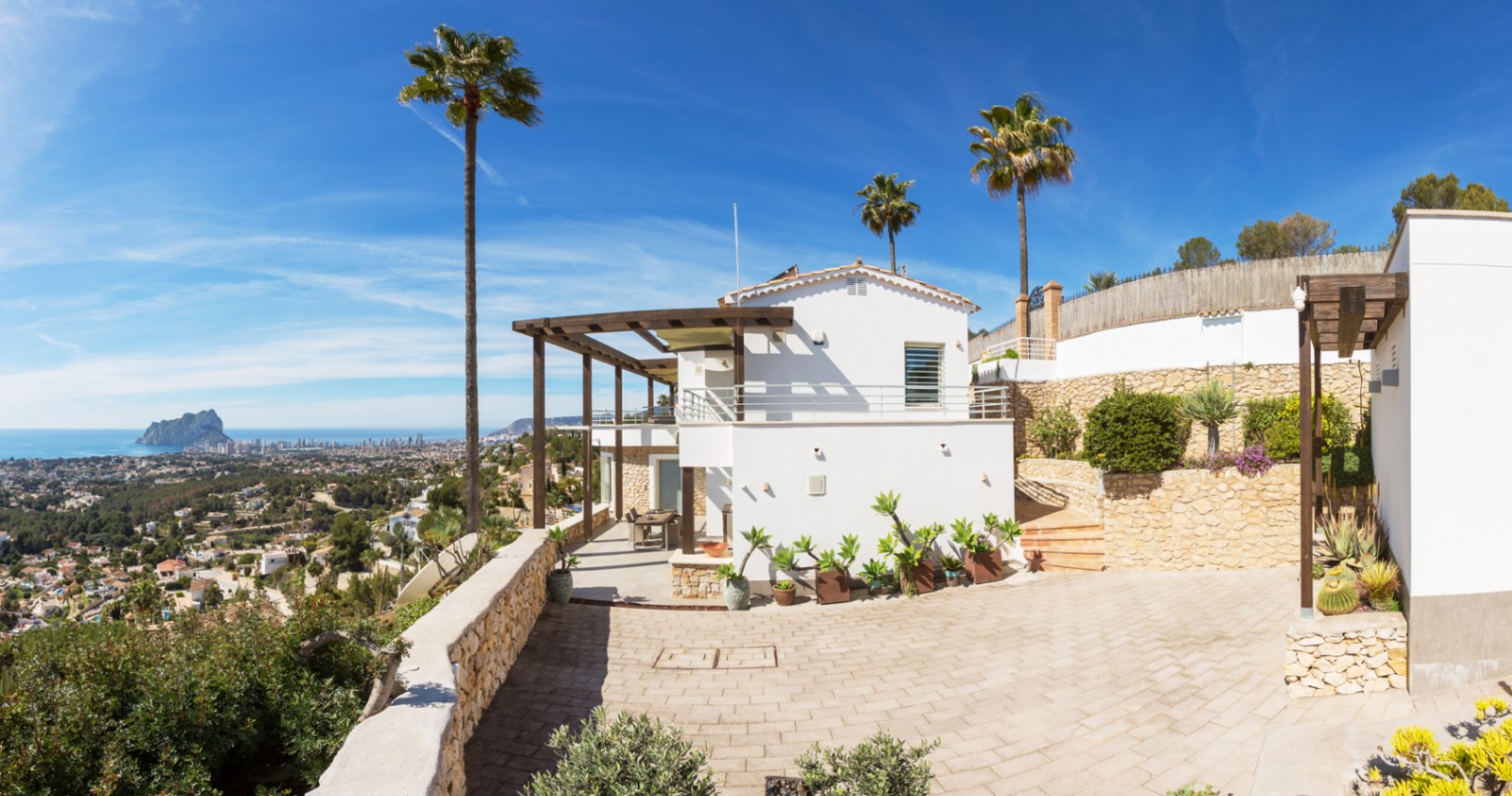 Exterieur van een mediterrane villa met witte gevels, houten pergola's en stenen muren, met panoramisch uitzicht op de kust en de Peñón de Ifach.