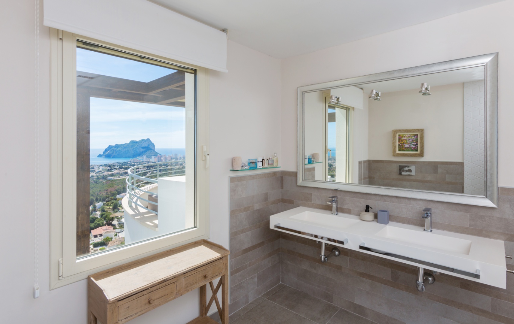 Moderne badkamer met dubbele zwevende wastafel, grote spiegel met zilveren lijst en raam met panoramisch uitzicht op de Peñón de Ifach en de kustlijn.