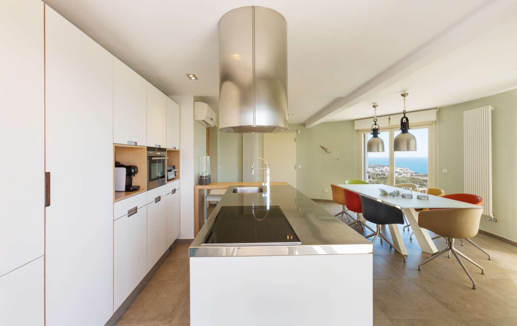 Moderne open keuken met wit kookeiland, roestvrijstalen werkblad, cilindrische afzuigkap en eethoek met uitzicht op zee door grote glazen deuren.
