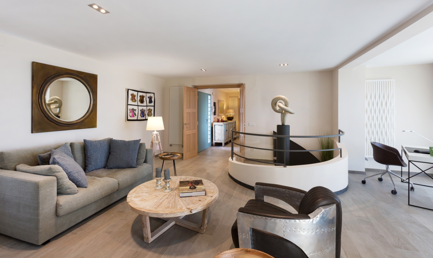 Moderne woonkamer met grijze bank, aviator-stijl fauteuil en een ronde trap met centraal beeldhouwwerk. Voorzien van werkplek en lichte houten vloer.