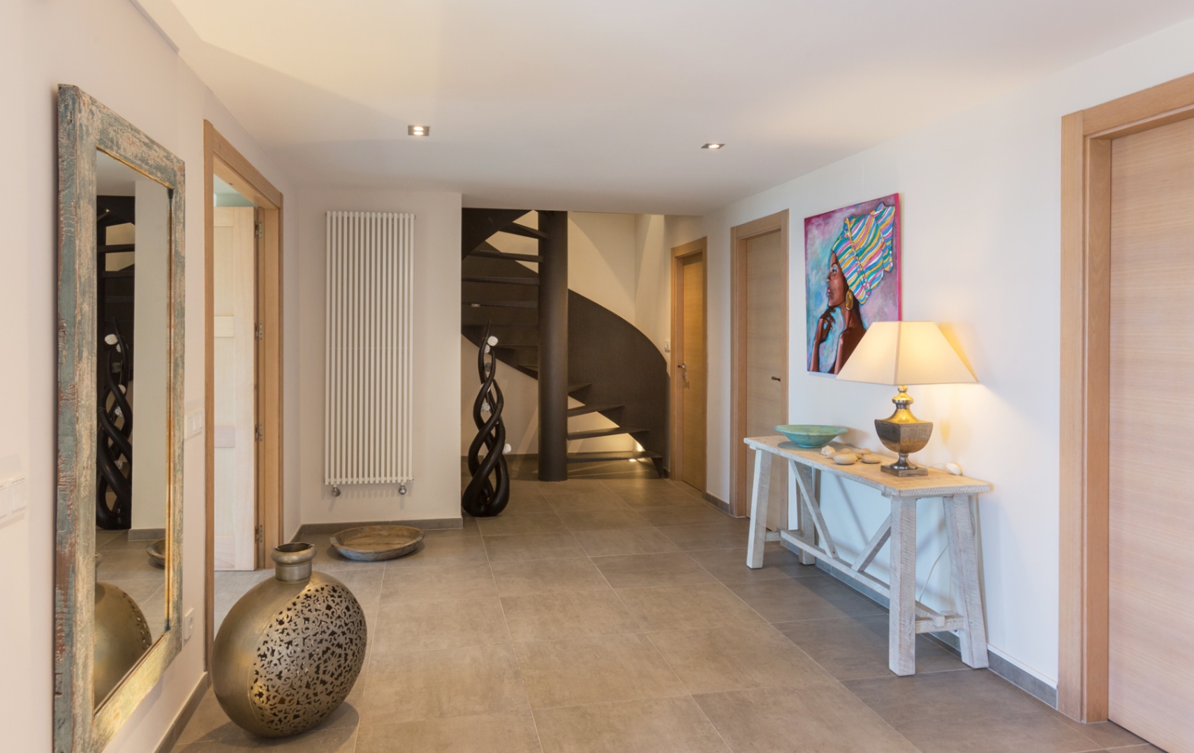 Ruime hal met grijze tegelvloer, een donkere metalen wenteltrap, licht houten deuren en een rustieke consoletafel met een decoratieve lamp en kunst.