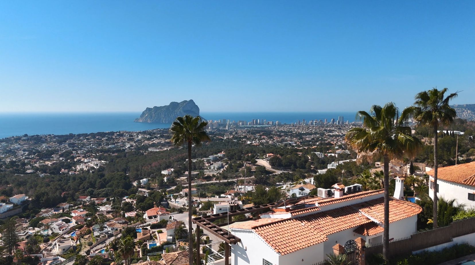 Verhoogd panoramisch uitzicht op de Peñón de Ifach en de skyline van Calpe, met mediterrane villa's met terracotta daken en palmbomen op de voorgrond.
