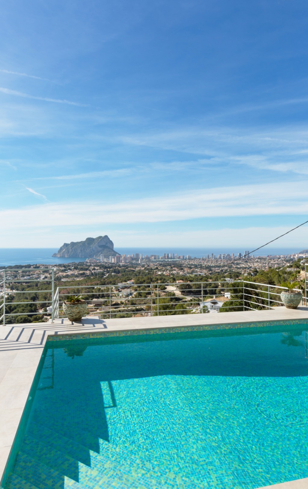 Verhoogd terras met een turquoise mozaïekzwembad en witte stenen rand, met panoramisch uitzicht op de Peñón de Ifach en de Middellandse Zeekust.