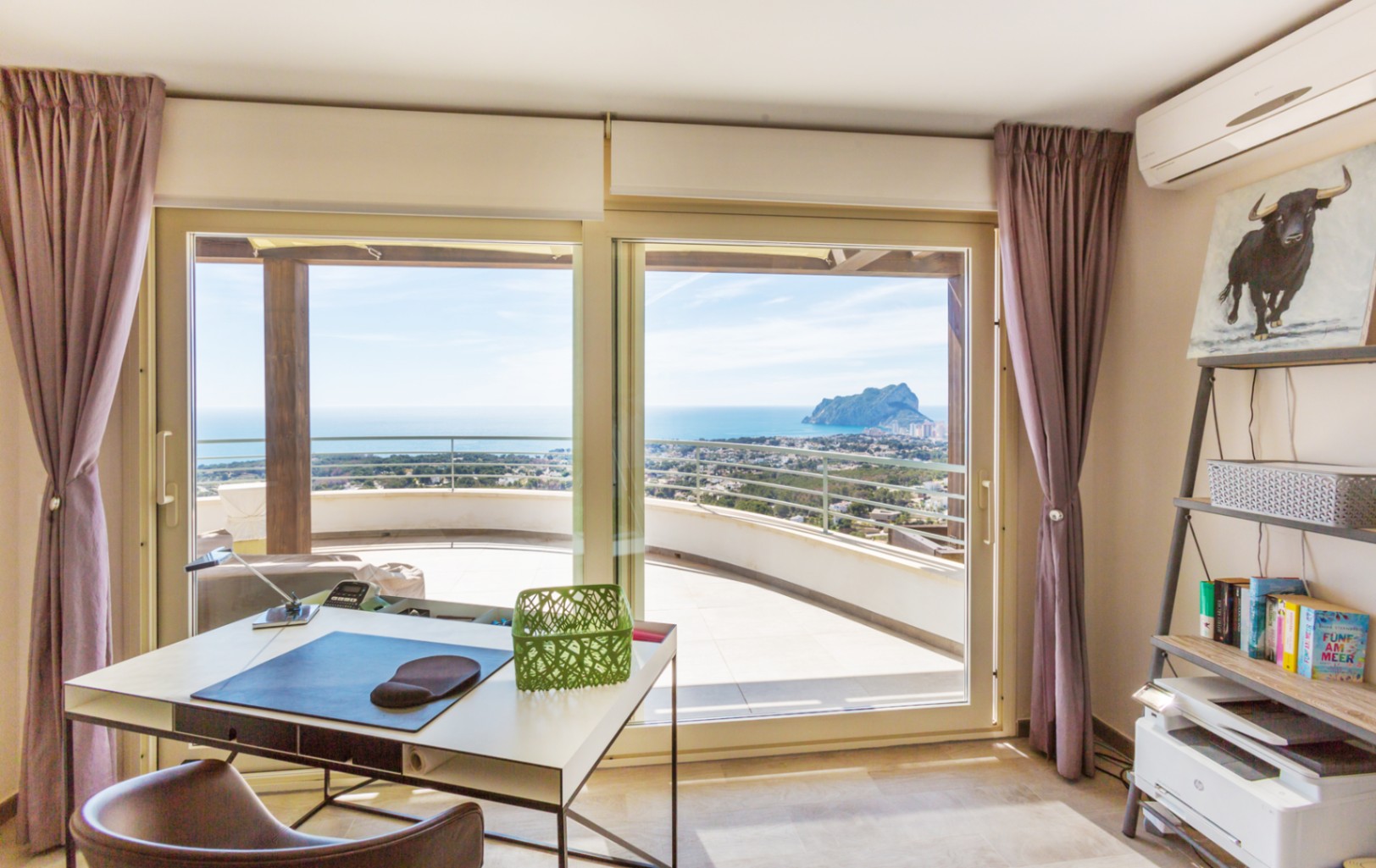 Werkkamer met modern bureau en stellingkast, voorzien van grote glazen schuifdeuren naar een terras met uitzicht op de Middellandse Zee en de Peñón de Ifach.