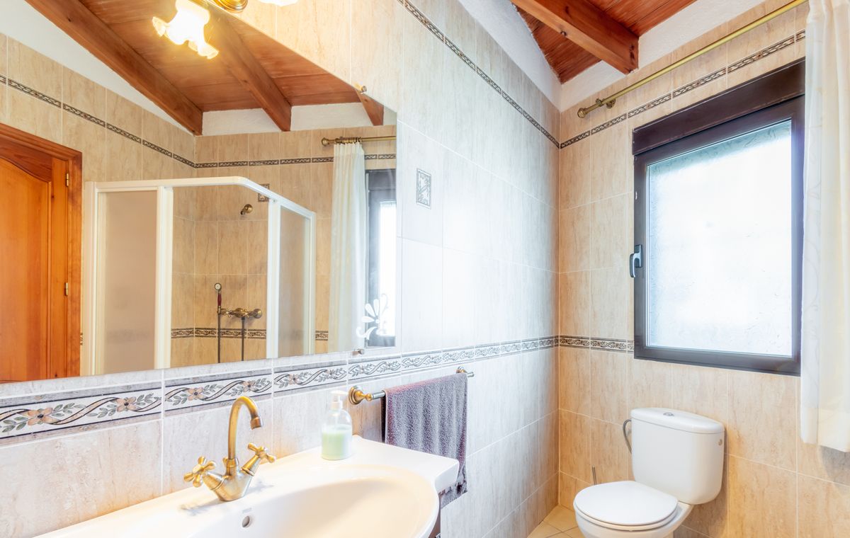 Badkamer met schuin houten plafond, zichtbare balken, beige tegels met sierranden en klassieke koperen kranen.