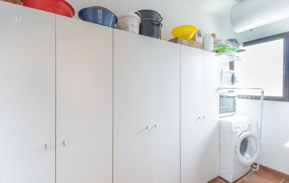 Bijkeuken met uitgebreide witte kastenwand, terracotta vloertegels en een wasruimte voorzien van een wasmachine en gestapelde apparatuur.