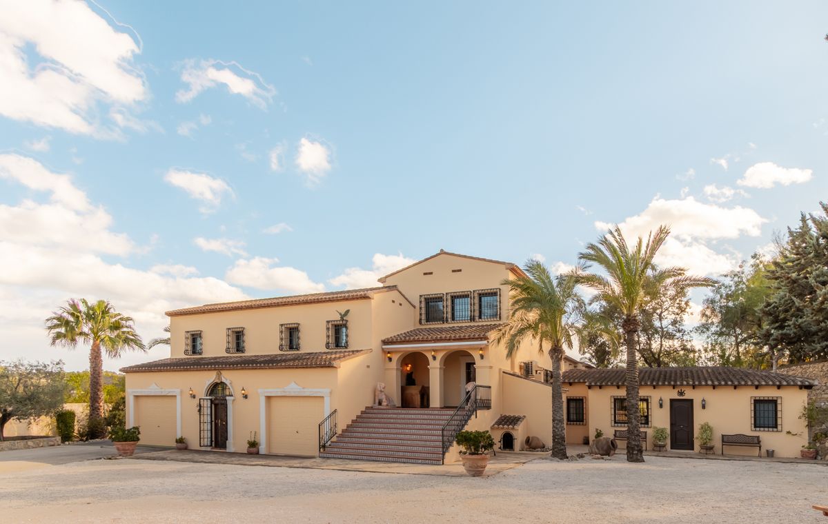 Exterieur van een mediterrane villa met crème gevel, terracotta dak, brede trap naar de boogingang en geïntegreerde garages tussen palmbomen.