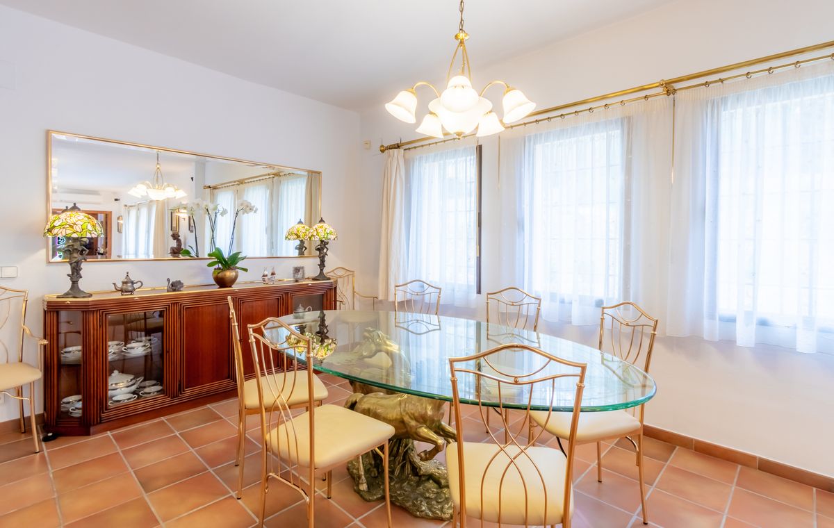 Formele eetkamer met een ovale glazen tafel op een sculpturale koperen voet, goudkleurige stoelen, terracotta vloer en een houten dressoir met spiegel.