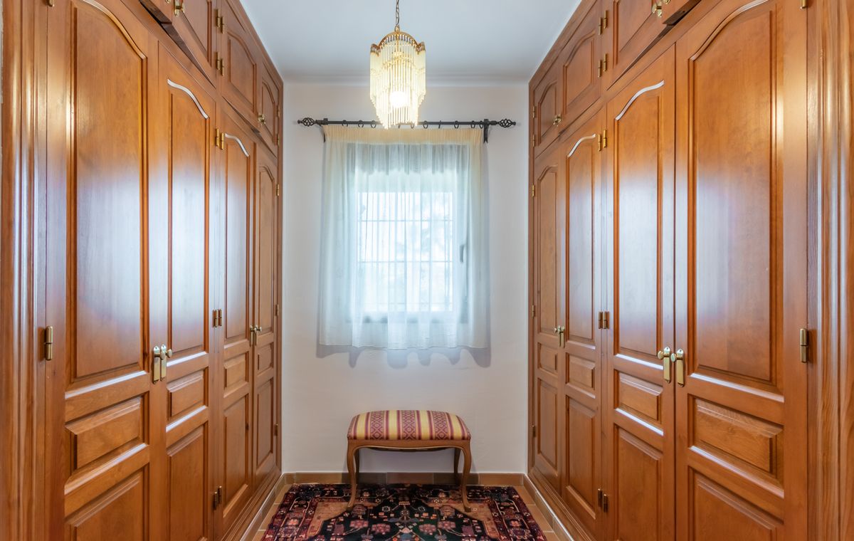 Inloopkast met kamerhoge houten inbouwkasten, een klassieke kroonluchter en een centraal raam voor natuurlijk licht.