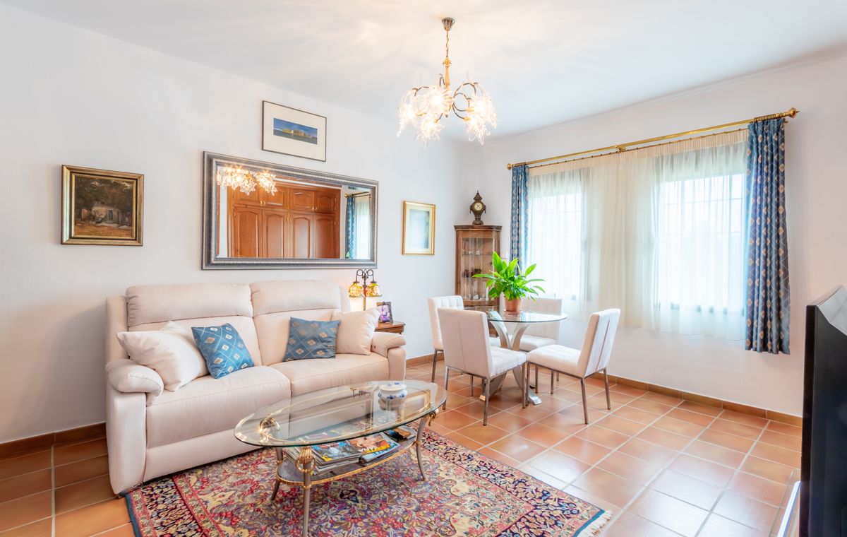 Lichte woonkamer met terracotta tegelvloer, een witte bank met blauwe kussens en een glazen eethoek onder een sierlijke glazen kroonluchter.