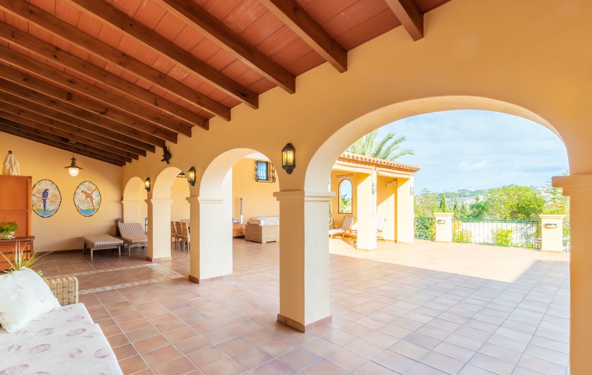 Overdekt terras met donkerhouten balken, terracotta tegelvloer met decoratieve randen en bogen die toegang bieden tot een groot open buitenterras.