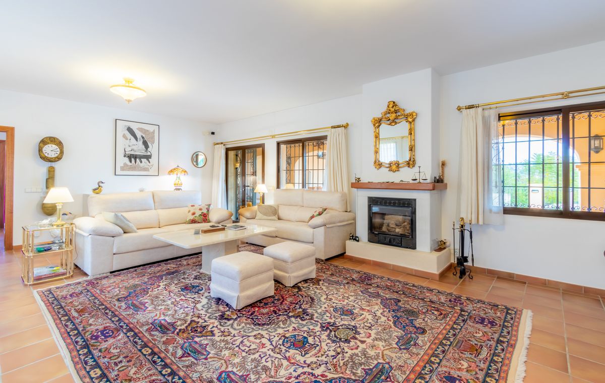 Ruime woonkamer met terracotta vloer, een klassieke haard met gouden spiegel en witte banken rondom een groot gedetailleerd tapijt.
