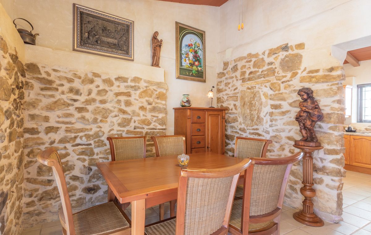 Rustieke eethoek met muren van natuursteen, een massief houten tafel met rieten stoelen en traditionele religieuze kunst en decoraties.