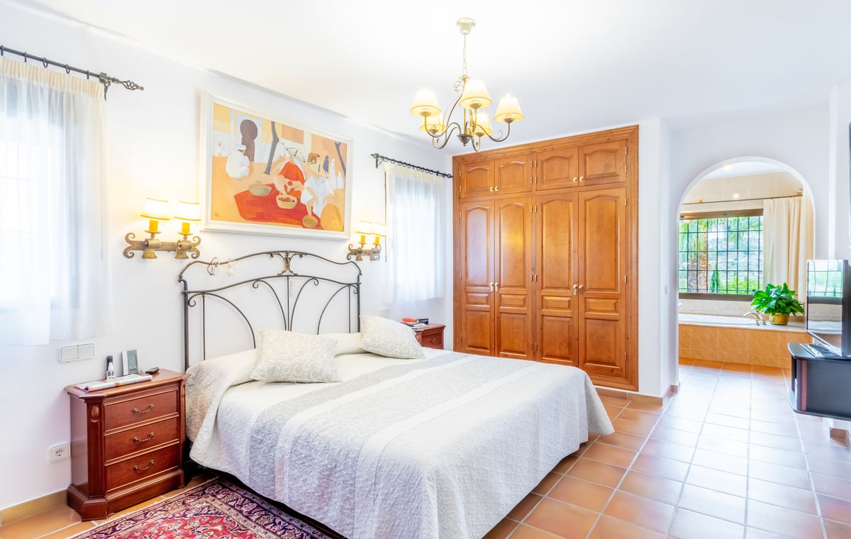 Slaapkamer met een smeedijzeren hoofdbord, houten inbouwkasten en een terracotta vloer. Een toog biedt toegang tot de aangrenzende badkamer met ligbad.