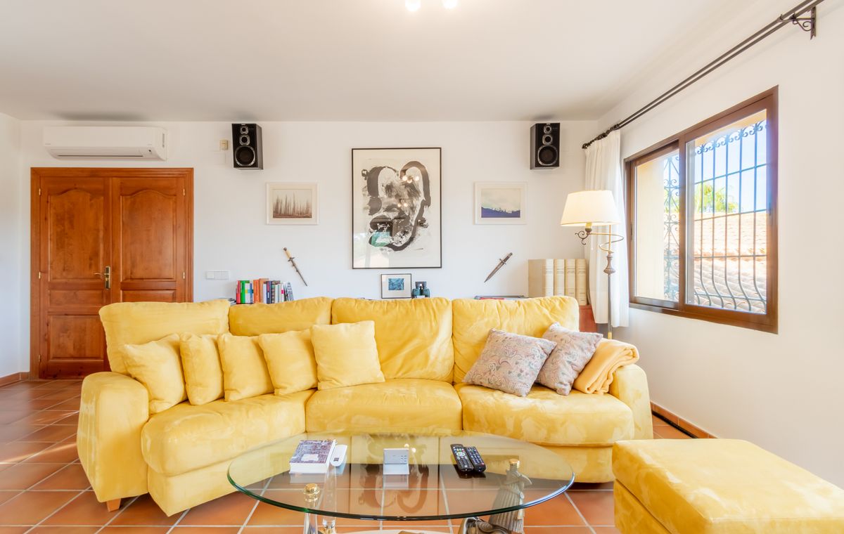 Woonkamer met een grote gele hoekbank, glazen salontafel en terracotta tegelvloer. De witte muren zijn gedecoreerd met kunstwerken en ingebouwde luidsprekers.